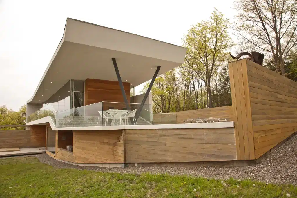 A striking modern home in Hudson, NY, showcasing a sleek design with glass railings, wooden accents, and an expansive outdoor area. The home features a unique angular roof and large windows, blending seamlessly with the surrounding trees and nature.