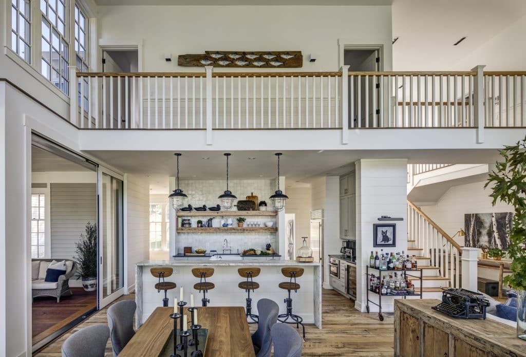 A bright, open-plan kitchen and dining area in a luxury custom home in the Hudson Valley featuring a wooden dining table with gray chairs in the foreground. Behind it, a kitchen island with wooden bar stools, industrial pendant lights, and open shelves with various decor items. Above, a balcony with white railings overlooks the space, and a cozy seating area is visible to the left.