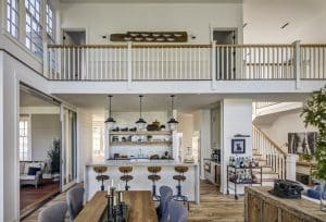 A bright, open-plan kitchen and dining area in a luxury custom home in the Hudson Valley featuring a wooden dining table with gray chairs in the foreground. Behind it, a kitchen island with wooden bar stools, industrial pendant lights, and open shelves with various decor items. Above, a balcony with white railings overlooks the space, and a cozy seating area is visible to the left.