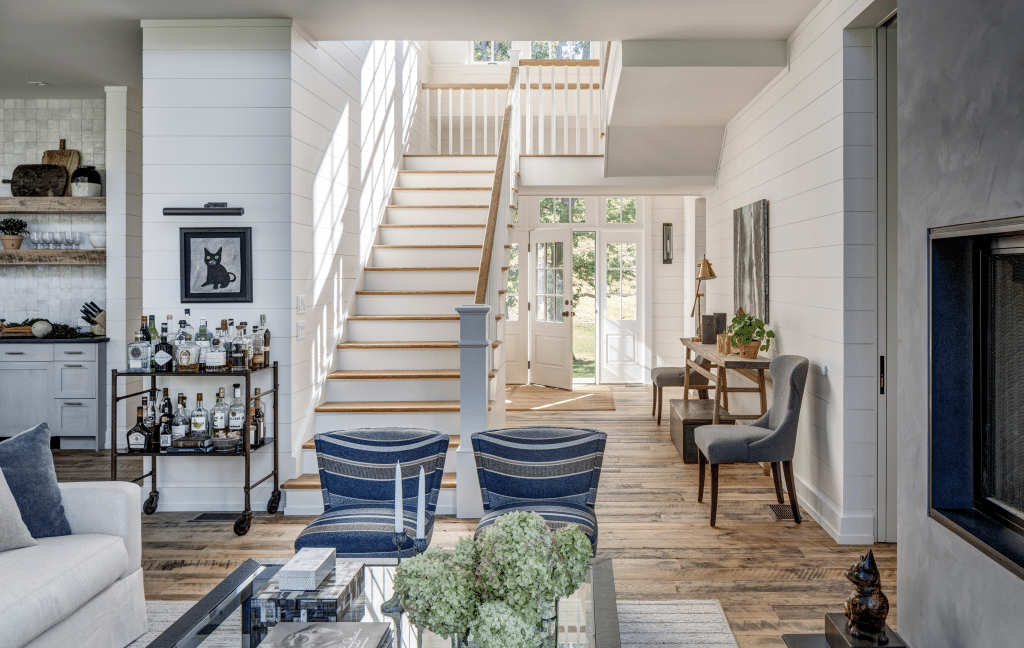 A spacious living room in a luxury custom home in the Upper Hudson Valley featuring a white staircase leading up to the second floor, large windows letting in abundant sunlight, and a well-stocked bar cart against a white shiplap wall. The room includes cozy seating areas and decorative elements like a black cat picture on the wall.