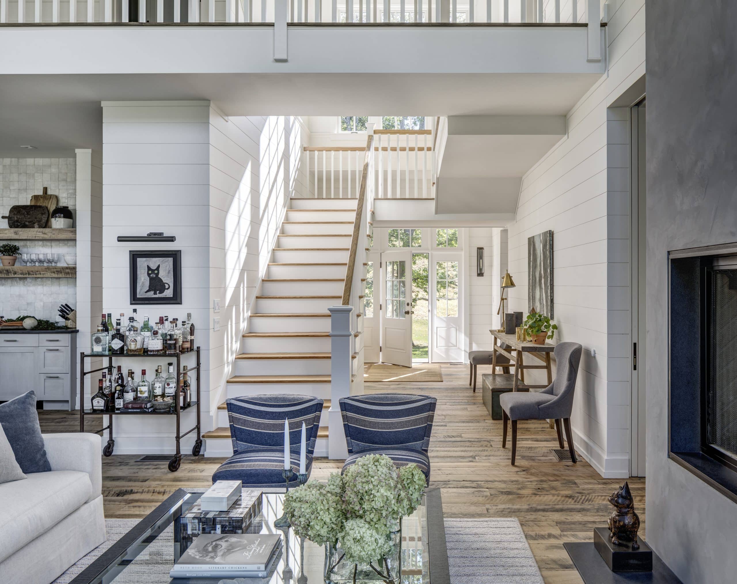 A spacious living room in a luxury custom home in the Upper Hudson Valley featuring a white staircase leading up to the second floor, large windows letting in abundant sunlight, and a well-stocked bar cart against a white shiplap wall. The room includes cozy seating areas and decorative elements like a black cat picture on the wall.