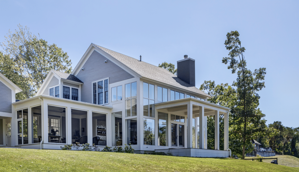 A charming gray house in the Hudson Valley, a luxury custom home with large windows and a glass-enclosed patio area, set against a backdrop of trees and a clear blue sky. The patio offers a panoramic view of the surrounding nature, providing a perfect space for relaxation and entertaining.