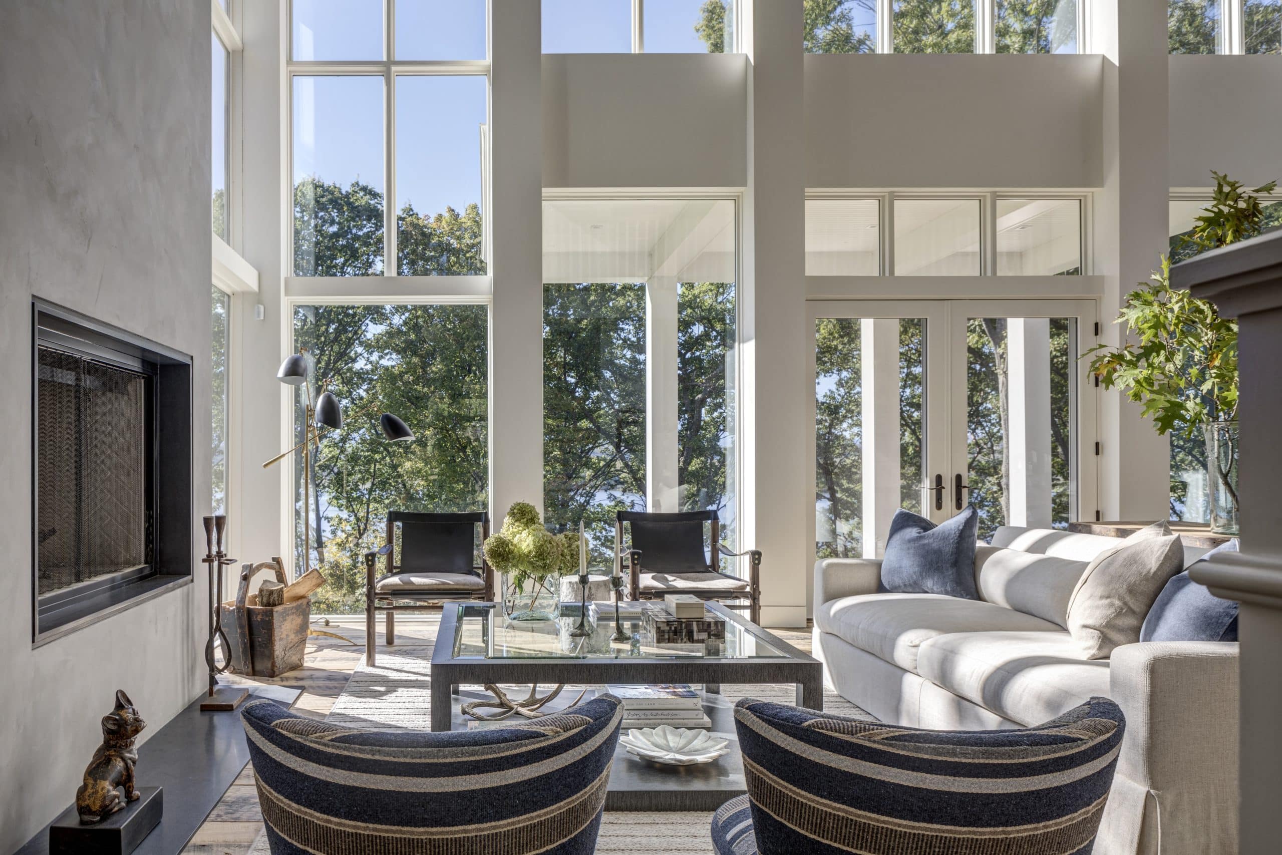 A beautifully designed living room in a luxury custom home in Hudson, NY with floor-to-ceiling windows providing ample natural light and stunning outdoor views. The space includes a comfortable beige sofa, blue-striped chairs, a glass coffee table, and modern decor elements like a black floor lamp and a fireplace.