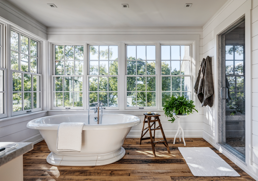 A luxurious bathroom in a luxury custom home in the Hudson Valley with a white freestanding tub positioned in front of large windows that offer a view of the surrounding greenery. The room features rustic wooden floors, white shiplap walls, and a small wooden stool beside the tub. A glass-enclosed shower is visible in the background.