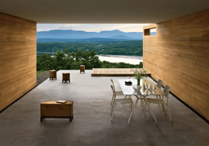 A minimalist outdoor dining space in a luxury modern home in the Upper Hudson Valley, featuring a simple glass dining table with white chairs. The area offers stunning views of the Hudson River and nearby mountains, surrounded by sleek wooden walls and an open concrete floor.