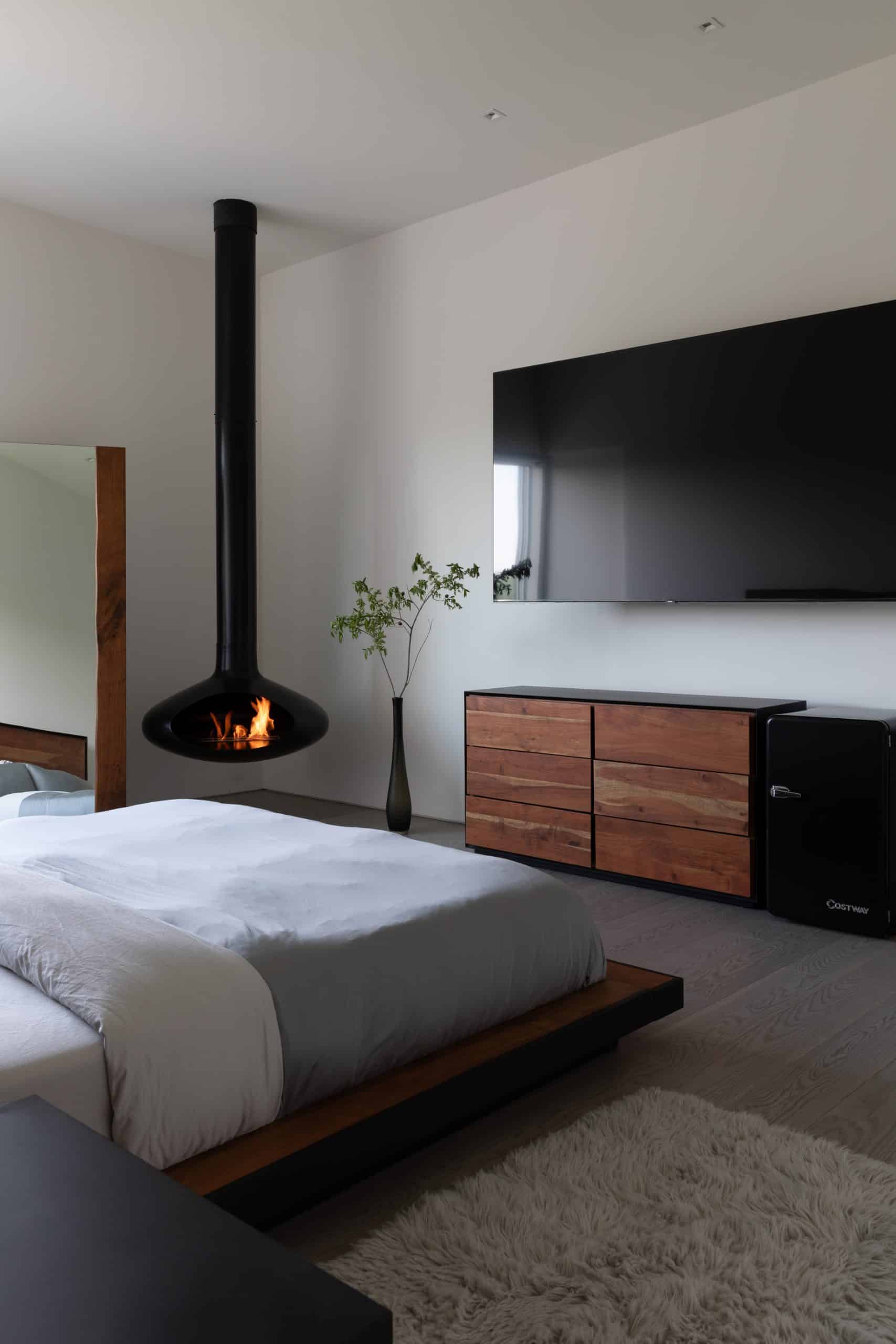 A sleek bedroom in a custom luxury home in the Hudson Valley, featuring a modern black hanging fireplace, a large bed with a gray duvet, a wooden dresser with natural wood tones, and a large wall-mounted TV. A tall vase with a green branch adds a touch of nature to the minimalist decor.