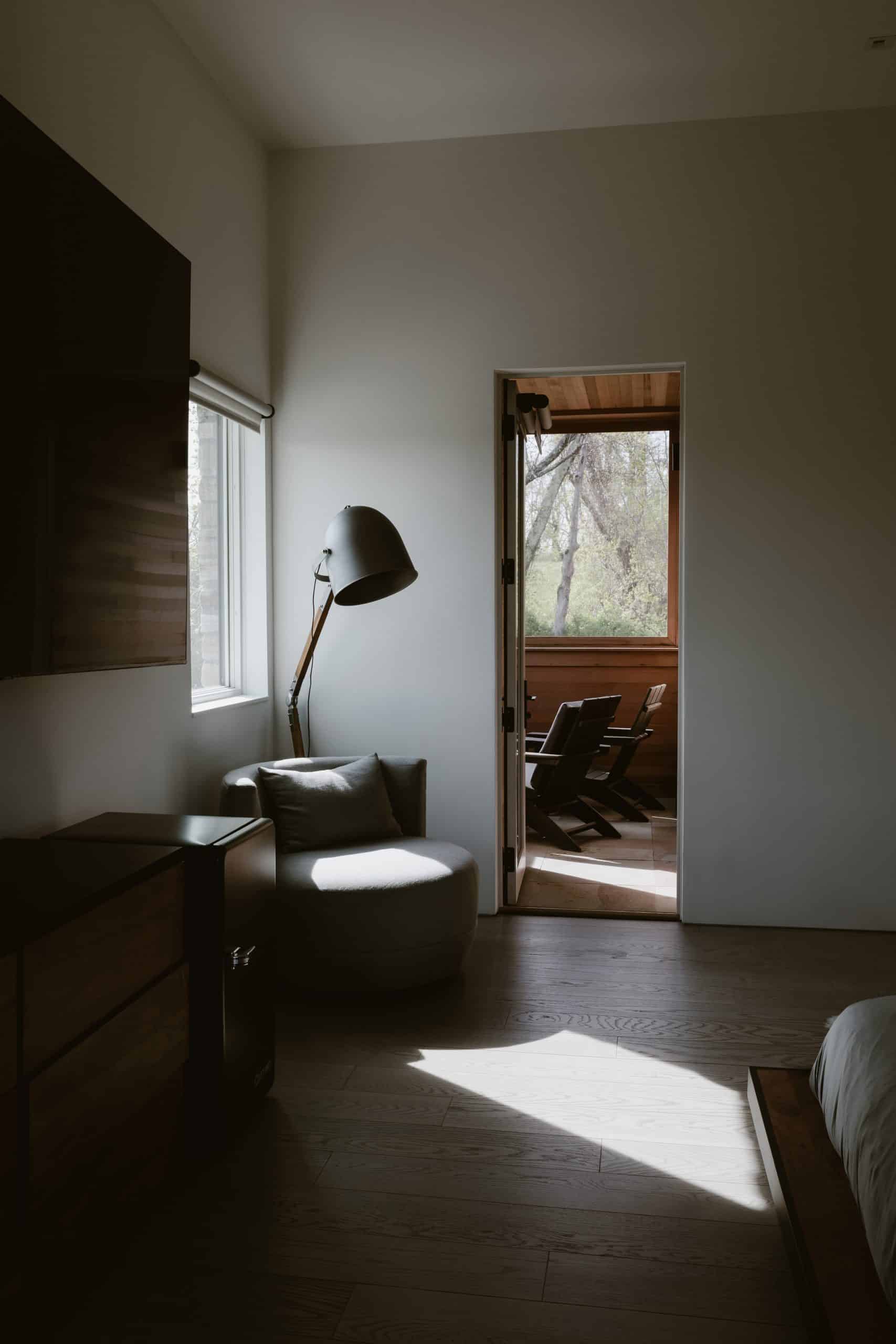 A stylish bedroom in a modern stone home in the Hudson Valley in Chatham, NY, featuring a cozy corner with a gray chair and a tall floor lamp. A window allows natural light to fill the space, while a doorway leads to an adjacent room with wooden paneling.