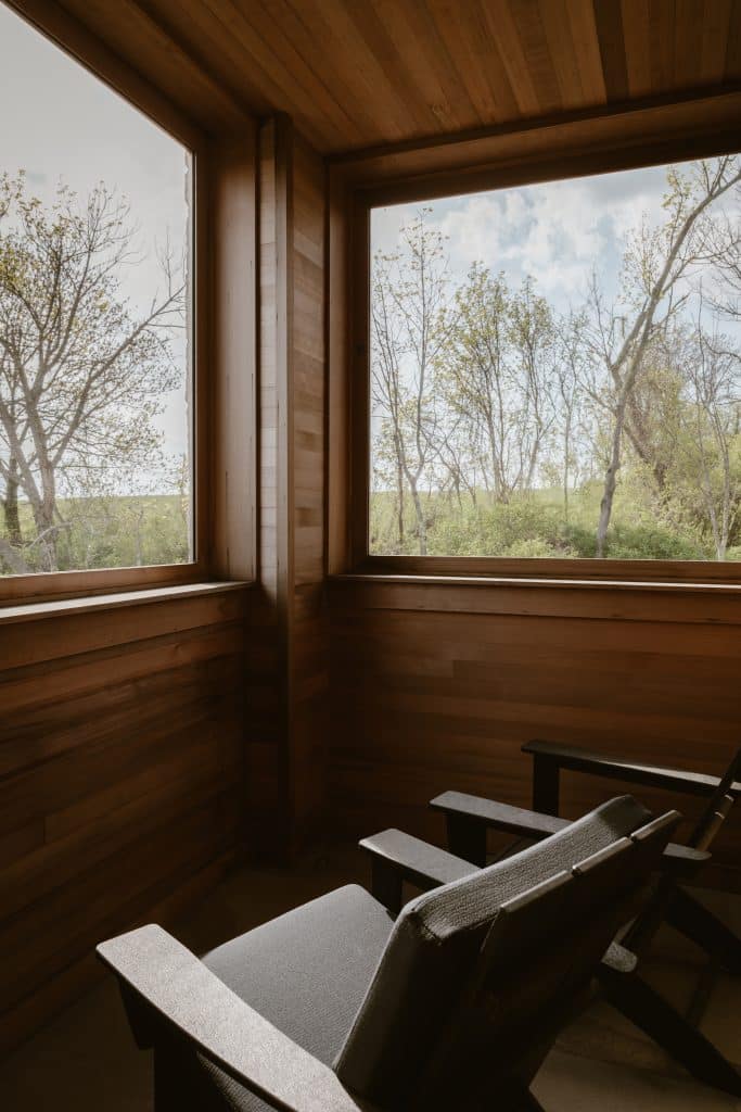 A cozy outdoor lounge in a custom stone cottage in the Upper Hudson Valley in Chatham, NY, featuring wood-paneled walls and ceiling. Large windows frame beautiful views of the surrounding forest, and two comfortable lounge chairs invite relaxation in this serene space.