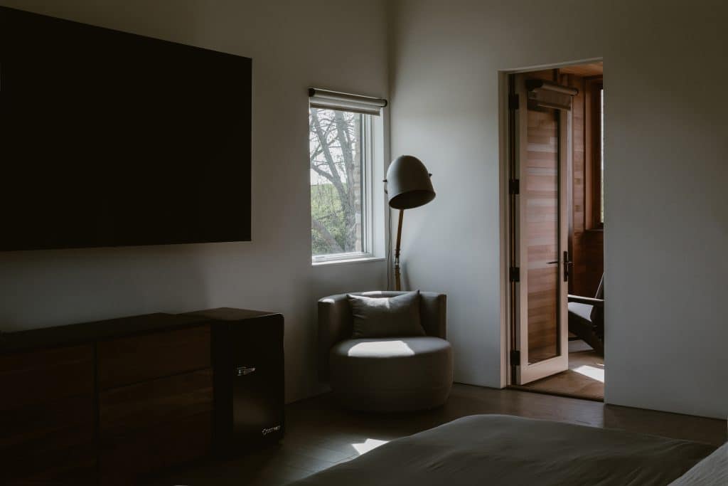 A tranquil bedroom in a custom luxury home in Chatham, NY, featuring a comfortable gray chair, a large floor lamp, and a doorway that leads to an outdoor lounge area with wooden chairs. The room is bathed in natural light from a nearby window, creating a peaceful atmosphere.