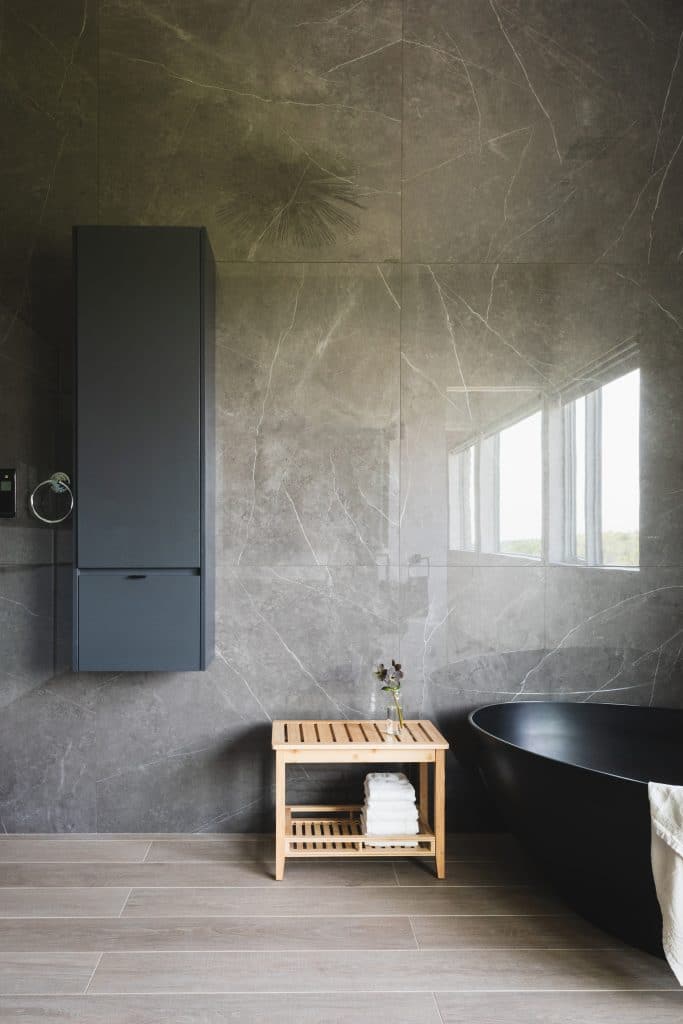 A modern bathroom in an Upstate New York custom stone cottage in Chatham, NY, featuring elegant stone walls, a sleek black bathtub, and minimalist wooden shelving. A small wooden bench with towels and a vase adds a touch of warmth to the space.