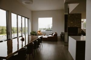 A spacious living and dining area in a modern stone home in Chatham, NY, featuring a large wooden dining table, leather couches, and floor-to-ceiling windows that offer views of the outdoor patio and surrounding nature.