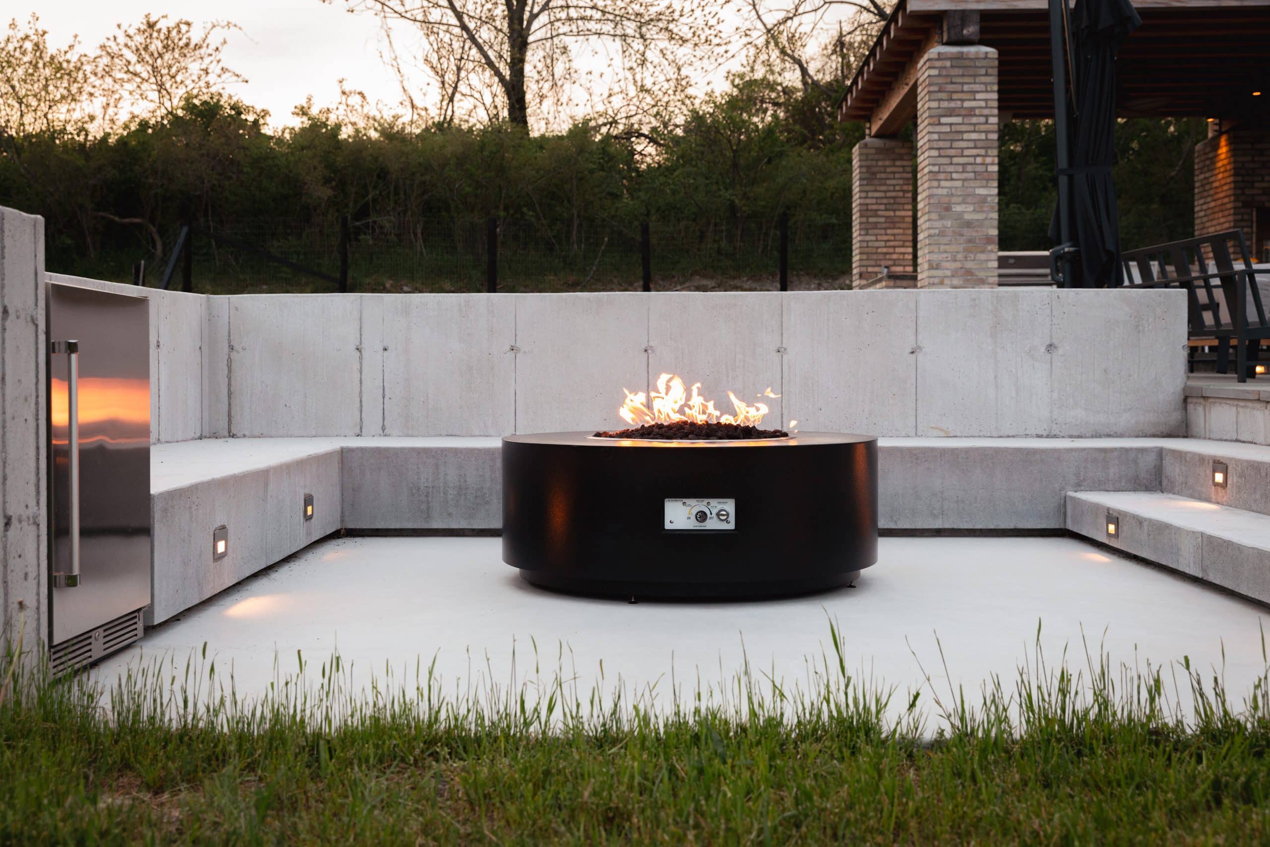 A modern outdoor fire pit area in a custom luxury home in Chatham, NY, featuring a sleek black fire pit, concrete seating, and ambient lighting set against a backdrop of trees and greenery.